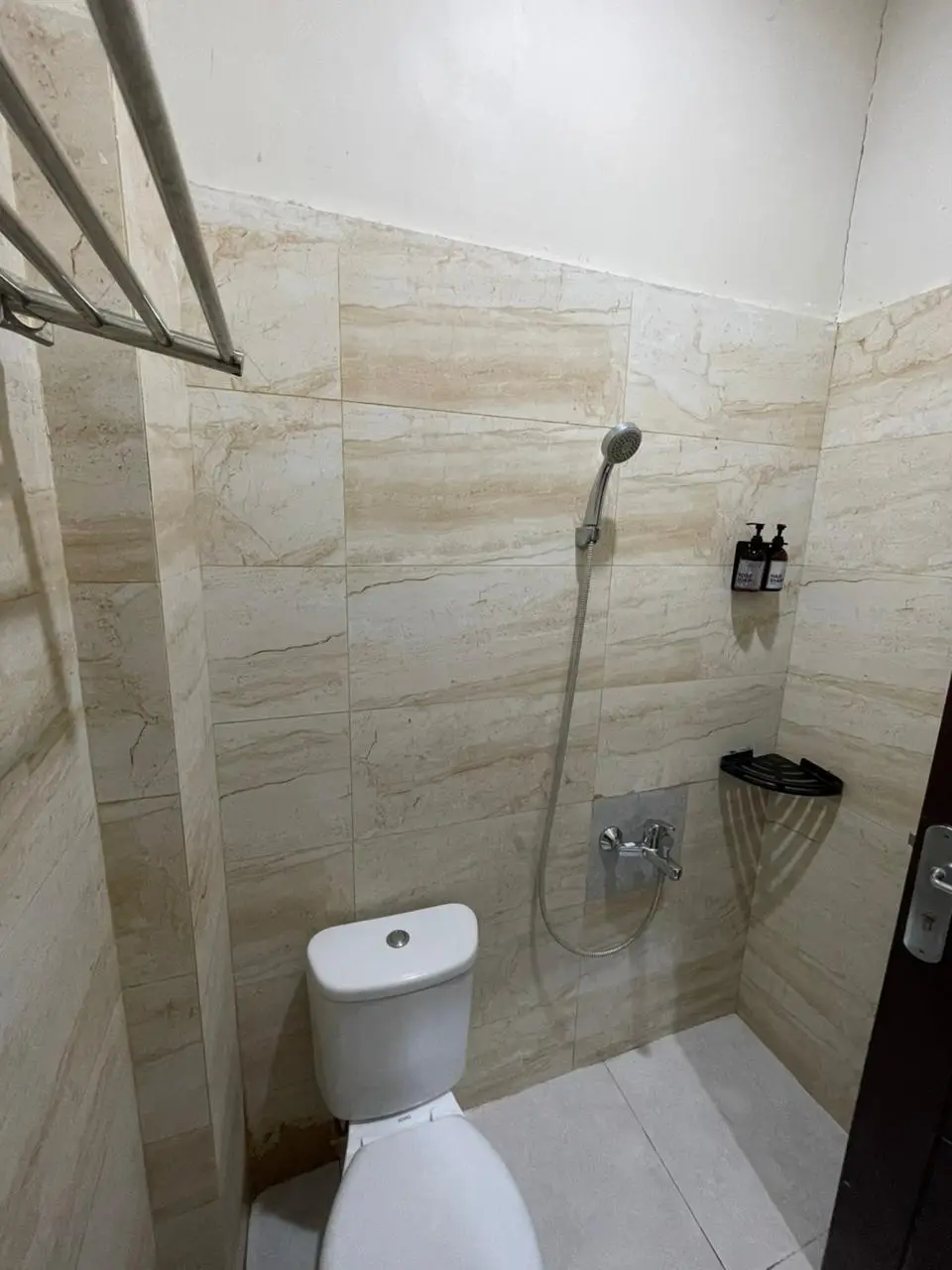 Kamar mandi hotel minimalis dengan dinding keramik marmer krem, toilet putih, dan shower.