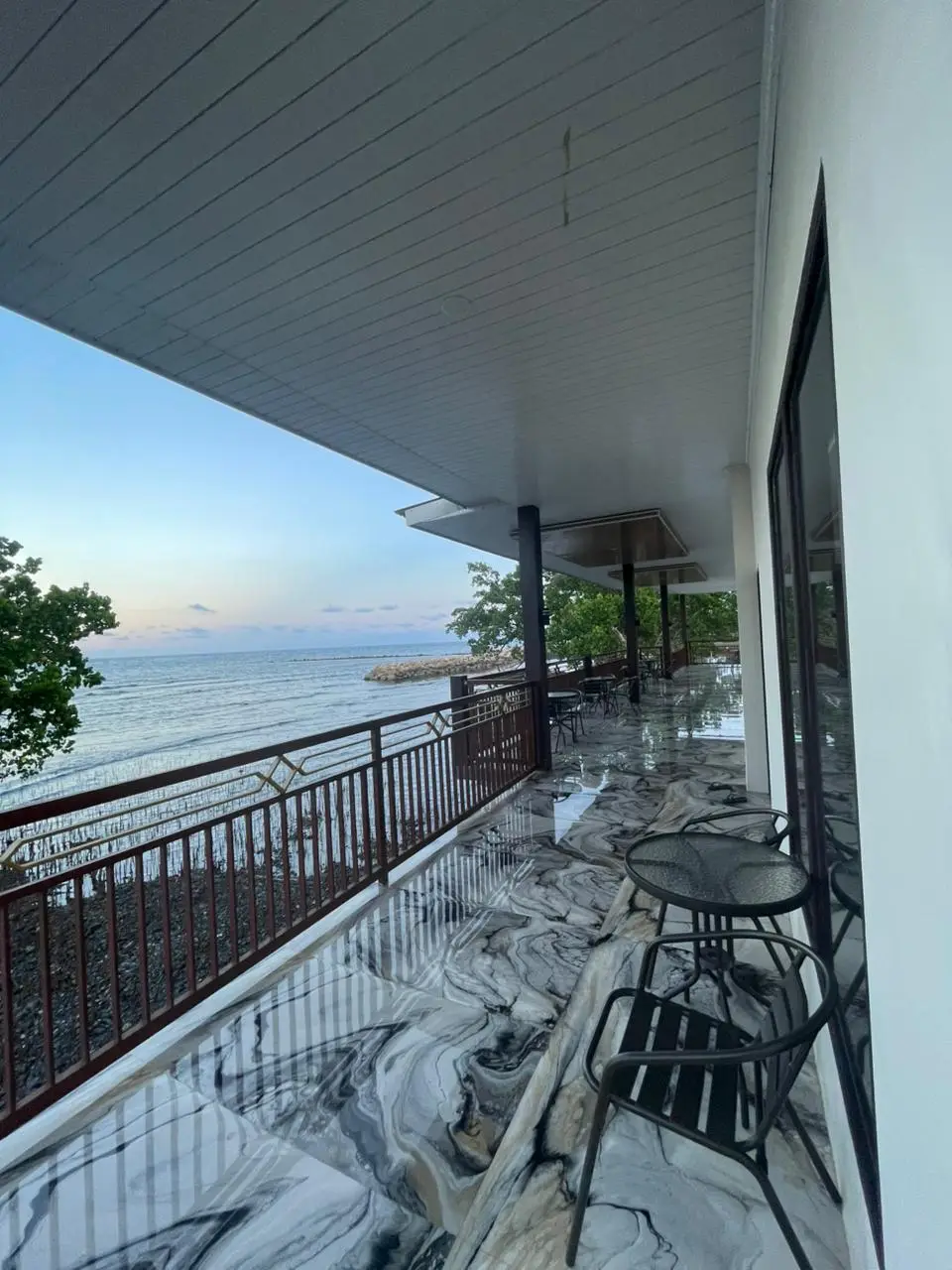 Balkon panjang kamar suite dengan lantai bermotif marmer menghadap ke laut saat matahari terbenam.