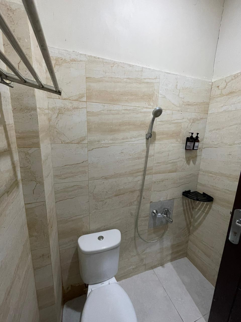 Kamar mandi hotel minimalis dengan dinding keramik marmer krem, toilet putih, dan shower.