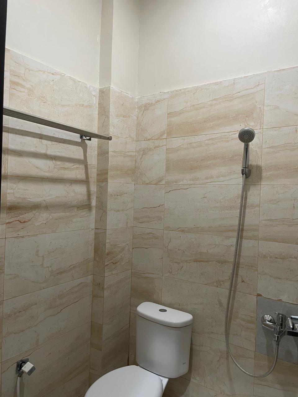 Tampilan dekat kamar mandi hotel dengan ubin dinding efek marmer, toilet putih, dan pancuran genggam perak.