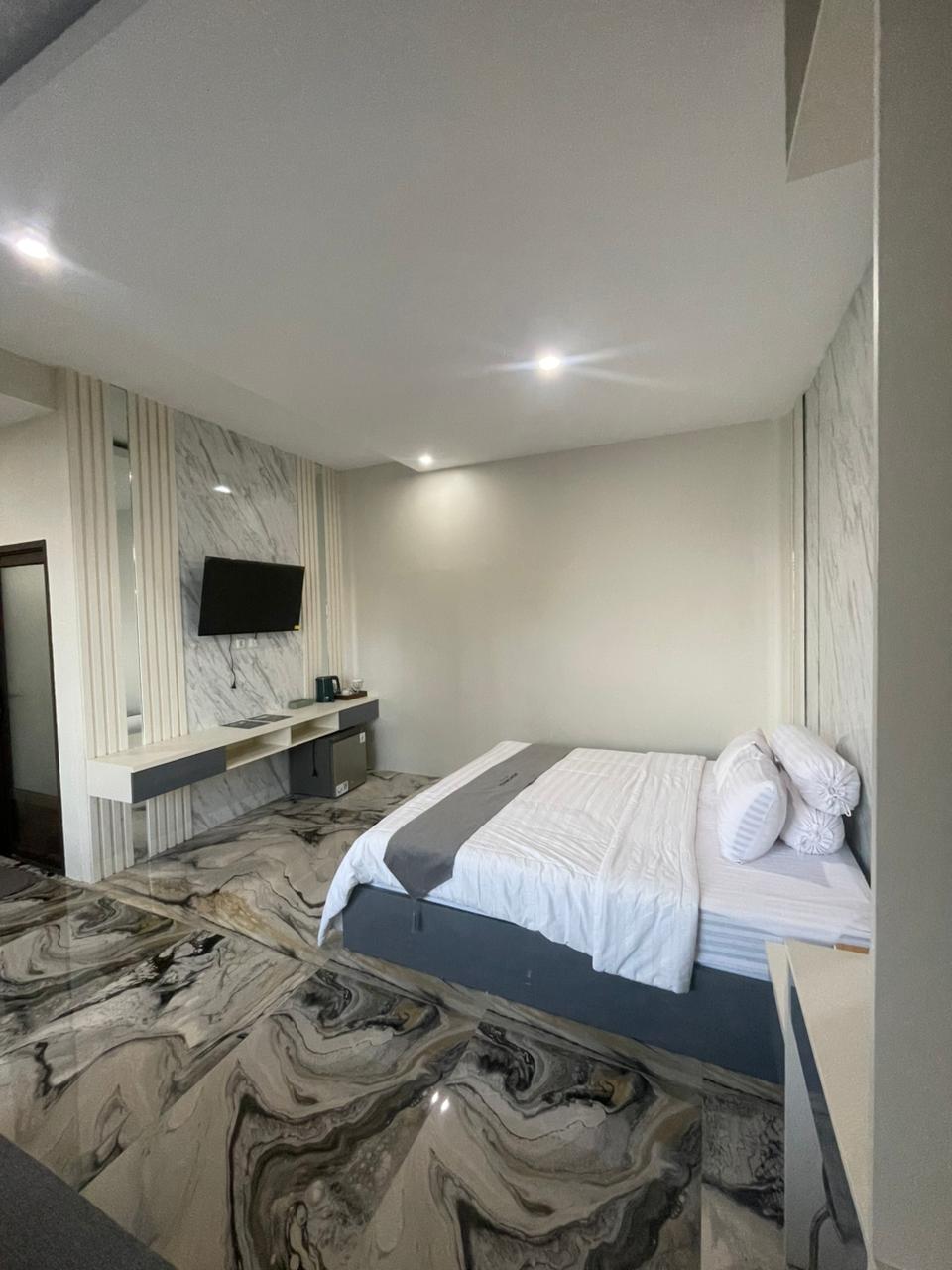 Kamar suite hotel modern dengan lantai bermotif marmer, tempat tidur king-size, dan televisi yang terpasang di dinding.